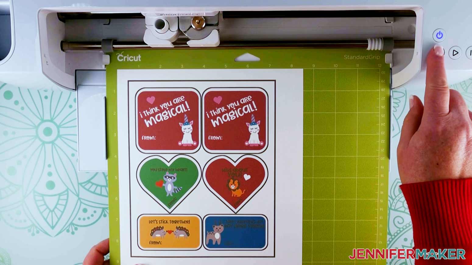 Closeup of a piece of white paper with colorful Valentine cards with cartoon animals stuck on a green machine mat and ready to be loaded and cut by a Cricut cutting machine.