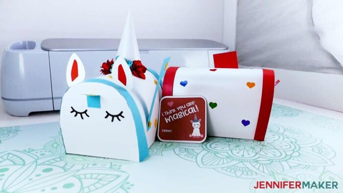 Two white paper Valentine Mailbox Crafts, one with blue accents that looks like a sleeping unicorn head next to a simpler version with red trim and various color hearts, and a red Valentine card with a unicorn saying "I think you are magical!" in front of a Cricut Maker.