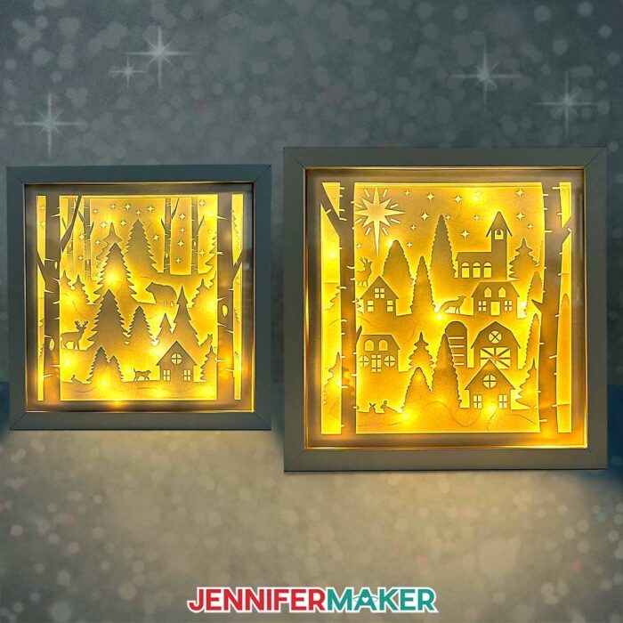 A winter forest shadow box with yellow lights and a village shadow box with yellow lights stand side by side.