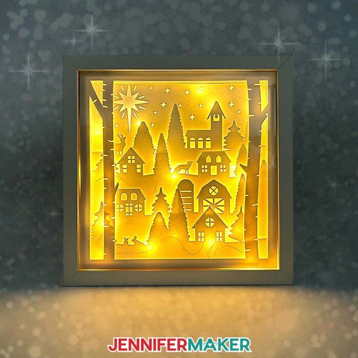 A Christmas scene houses, trees, and stars made from layered cardstock inside a shadow box lit with warm, yellow lights.