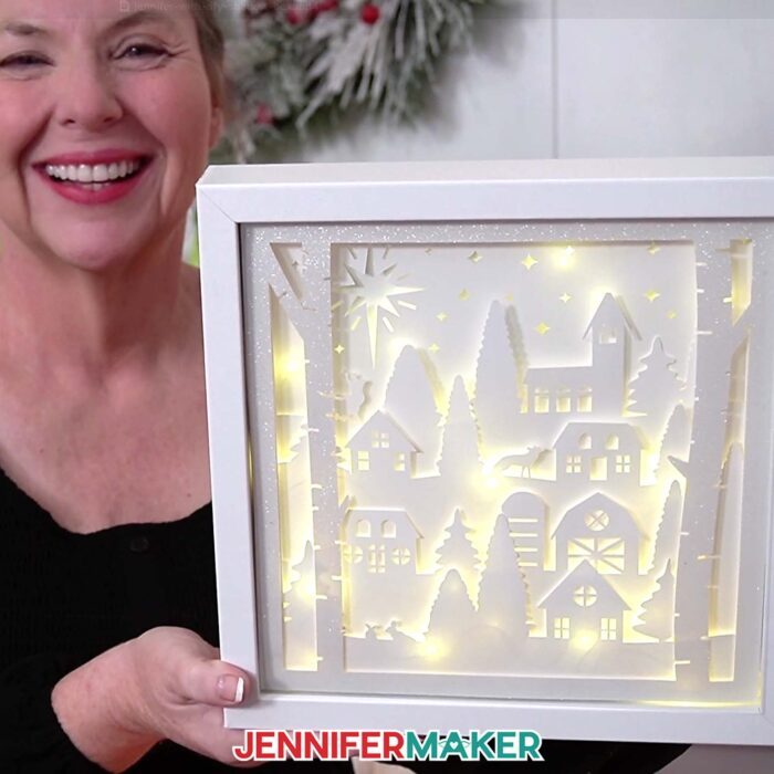 A close-up of Jennifer holding up the village shadow box with glowing lights behind the cardstock.