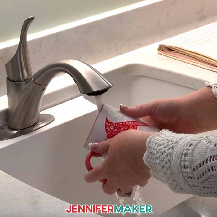 Hands hand-wash a white mug with a red cardinal adhesive vinyl design. Learn how to use Adhesive Vinyl Vs Iron-On For Cricut Beginners