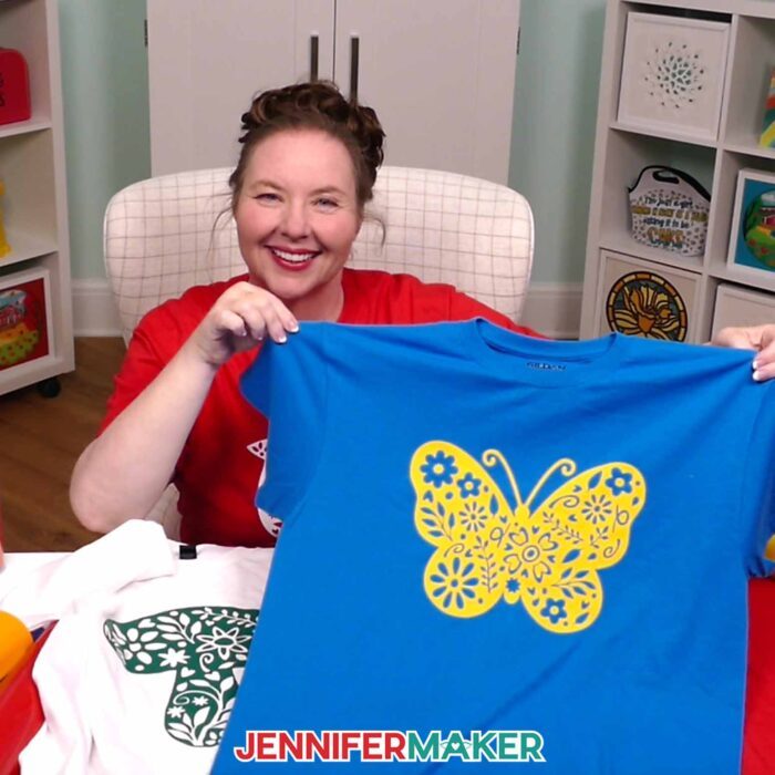 Jennifer Maker sits smiling at her craft table, showing off a blue T-shirt decorated with a yellow iron-on vinyl decal of a floral butterfly. Learn how to use Adhesive Vinyl Vs Iron-On For Cricut Beginners