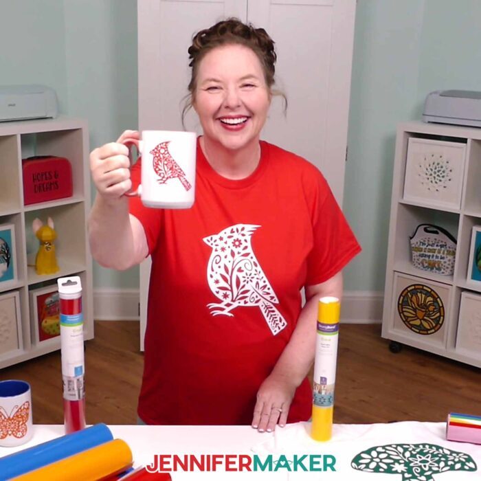 Jennifer Maker wears a red T-shirt decorated with a white floral cardinal iron-on vinyl design, and holds out a white mug decorated with a red floral cardinal design. Learn how to use Adhesive Vinyl Vs Iron-On For Cricut Beginners