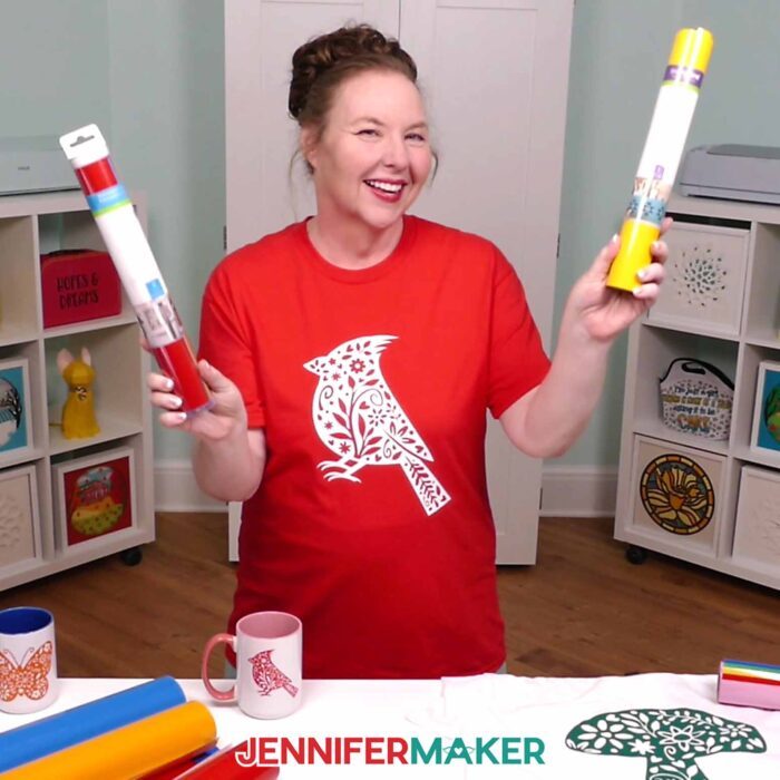 Jennifer Maker wears a red T-shirt decorated with a white floral cardinal iron-on vinyl design, holding out a white mug with a red floral cardinal adhesive vinyl design. Learn how to use Adhesive Vinyl Vs Iron-On For Cricut Beginners