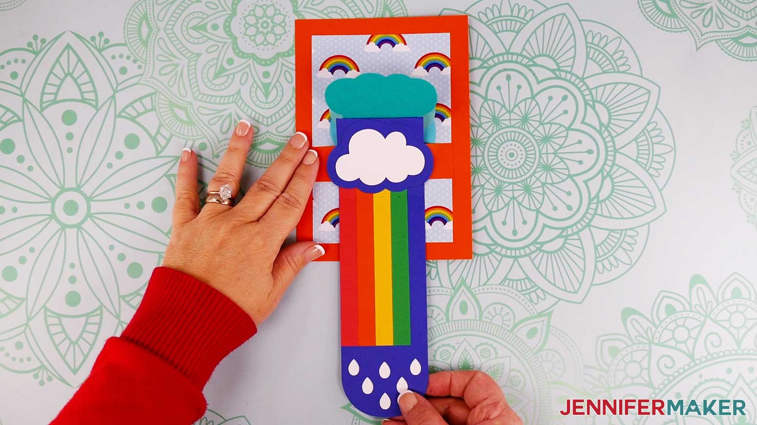 The assembled waterfall card is being opened by pulling down on the raindrop slider section, revealing the rainbow stripes beneath the cloud pieces.