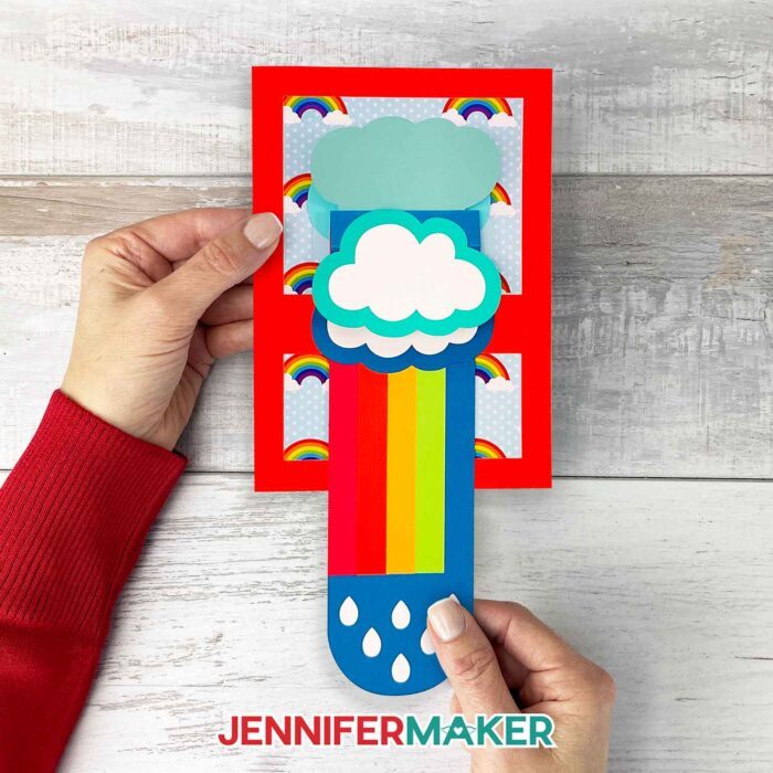 A person pulling the tab on a JenniferMaker waterfall card to reveal most of the rainbow and flip up the top three clouds.