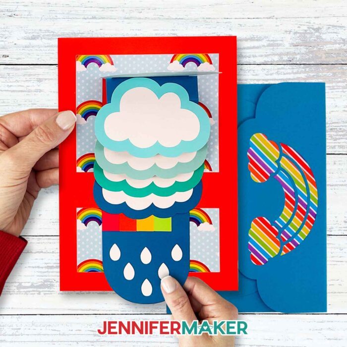 A person pulling the tab on a JenniferMaker waterfall card to reveal the rainbow and flip up the top cloud.