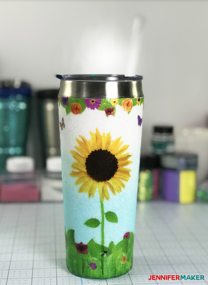 Drinking tumbler with an illustrated sunflower design on it in front of craft supplies.
