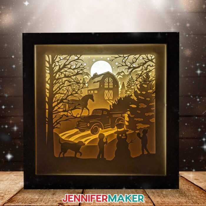 Learn to reinforce intricate paper cuts with vinyl in Jennifer Maker's winter shadow box tutorial! A glowing cardstock shadowbox featuring a winter scene with a barn, truck, horse, and children building a snowman (with the help of their dog) illuminates a dark room.