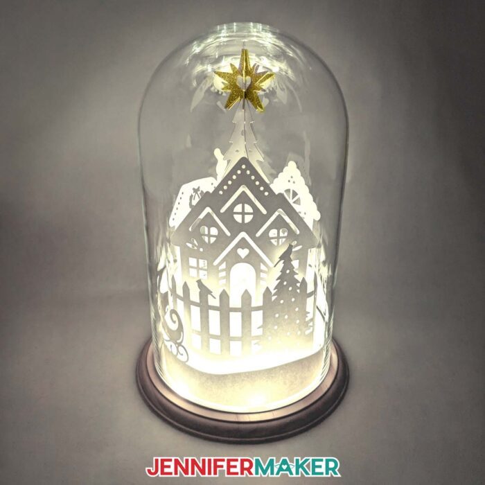 A 3D Cricut cut paper village lit up in a glass dome on a table.
