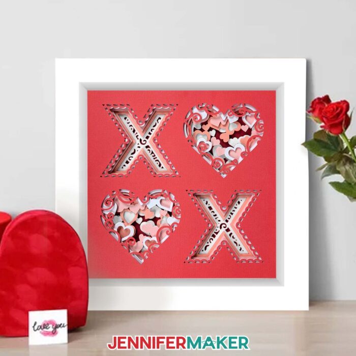 Learn to make a Love Shadow Box! A cardstock shadow box with a red X and O design with layers of red, pink, and white hearts sits on a table.
